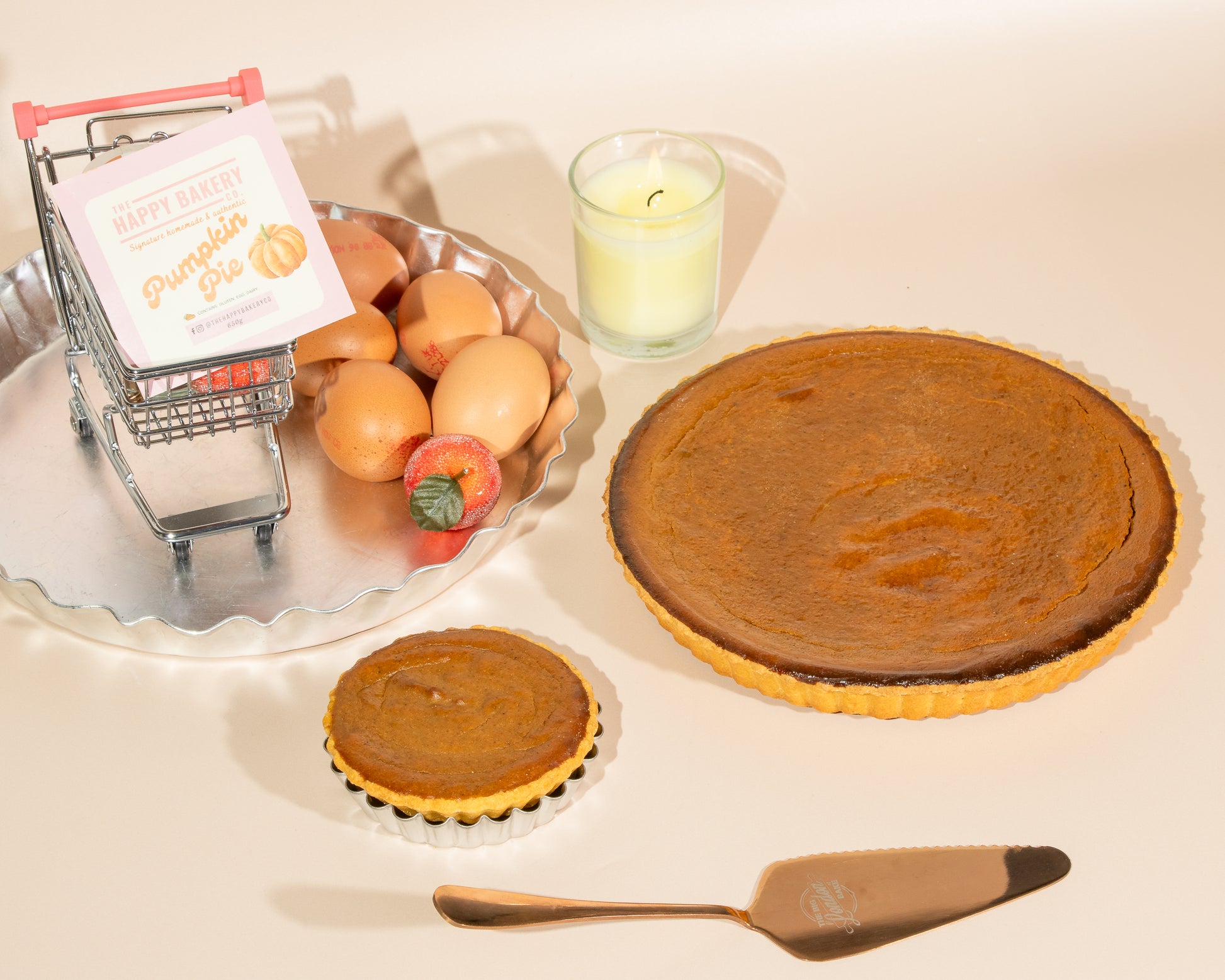 Pumpkin pies on a table with a small shopping cart, apples, and a candle.